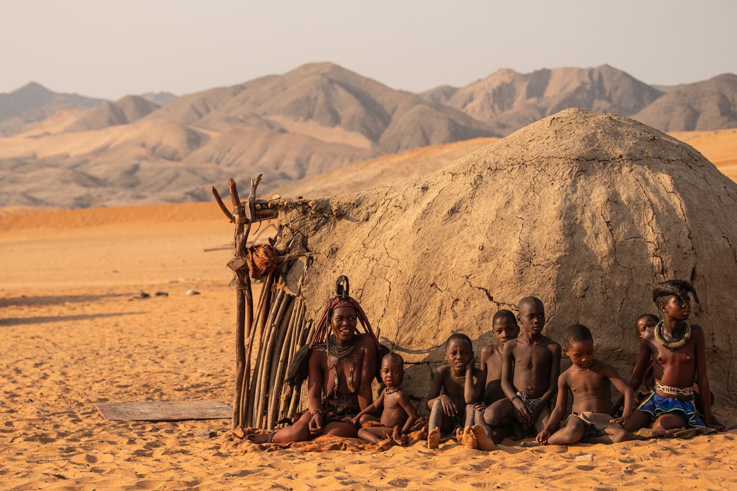 Himba tribe family sitting outside traditional mud hut in Namibian desert with mountain backdrop.