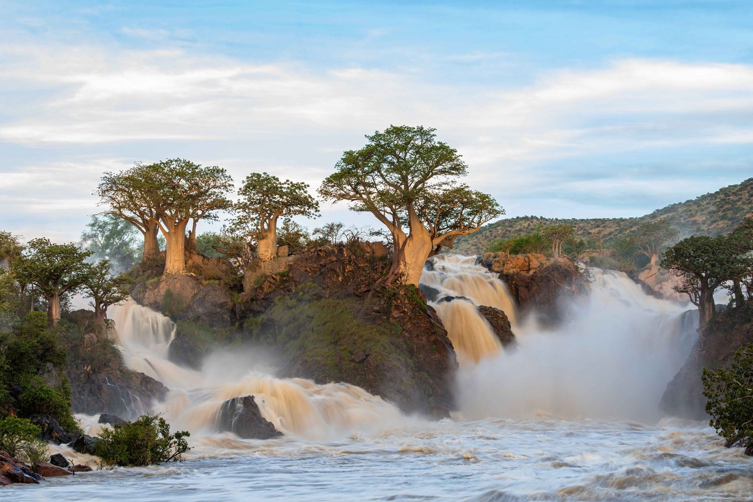 Rushing waterfalls cascade around rocky islands topped with baobab trees under a blue sky.