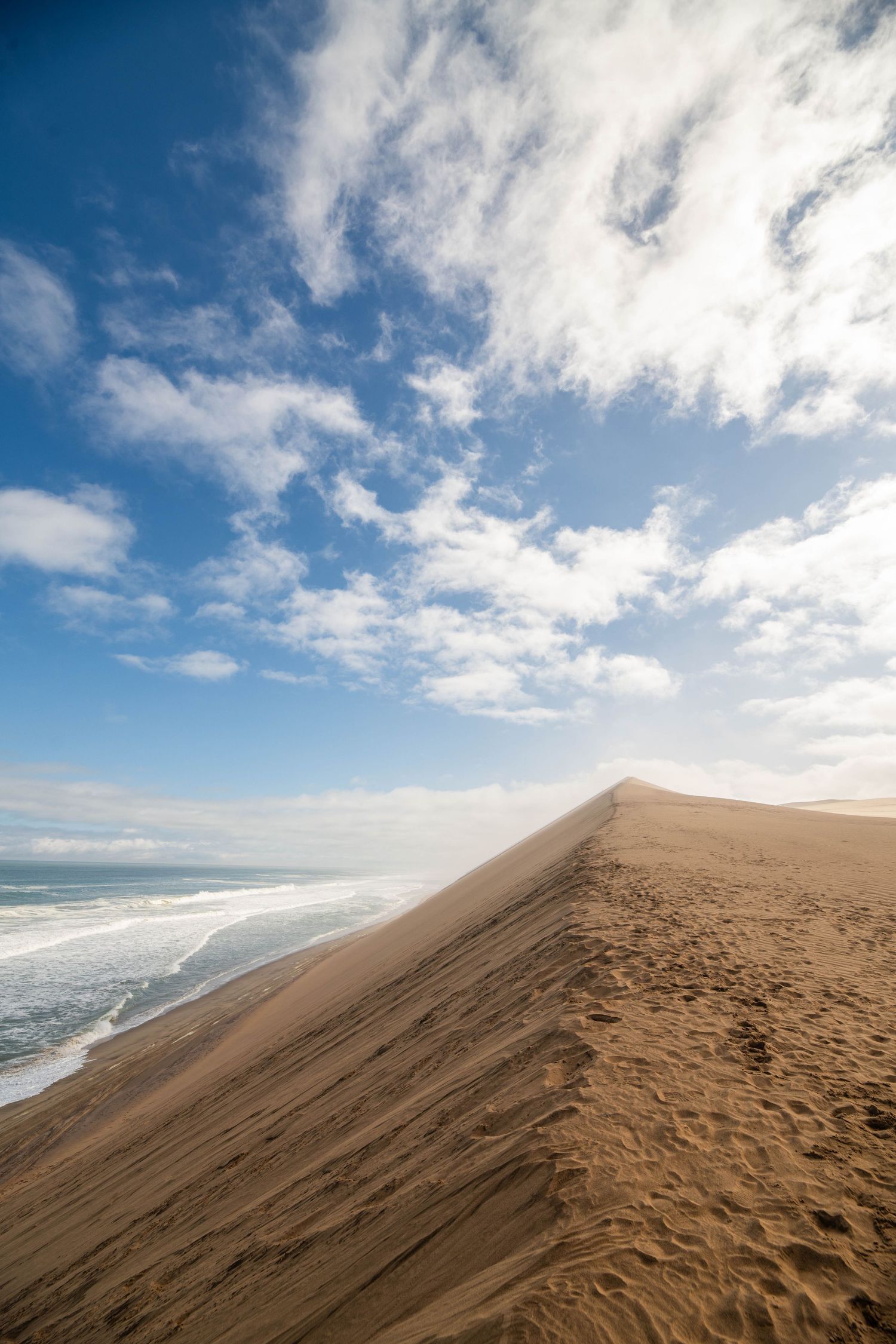 Steep sand dune ridge meeting Atlantic Ocean waves under blue sky with scattered white clouds.