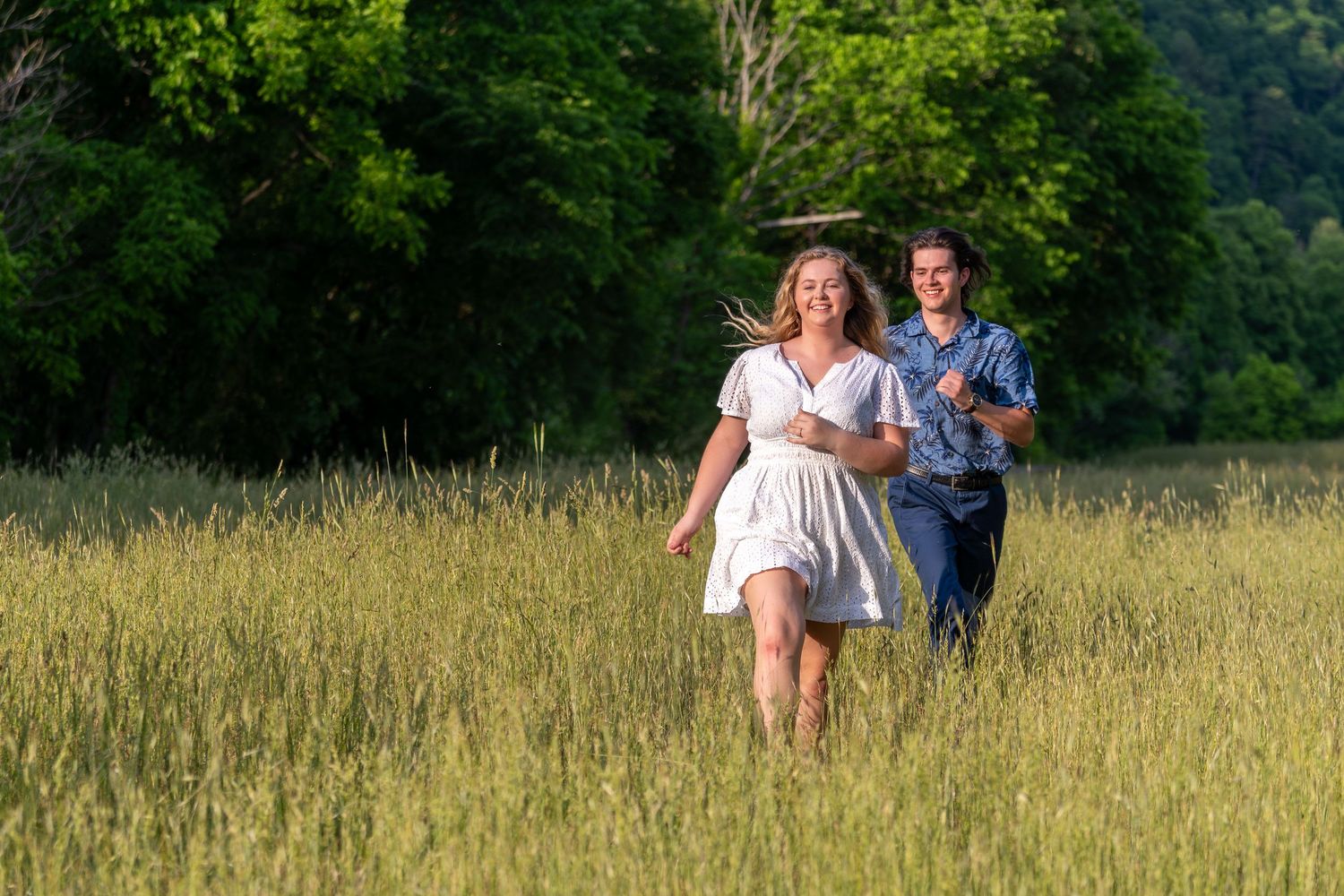 Couple running and laughing together through a sunlit green meadow with trees in background.
