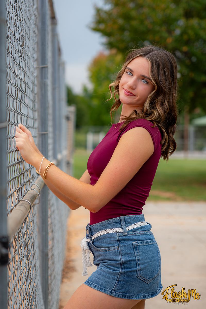 Young woman in maroon top and denim shorts leaning against a chain-link fence at an outdoor sports facility.
