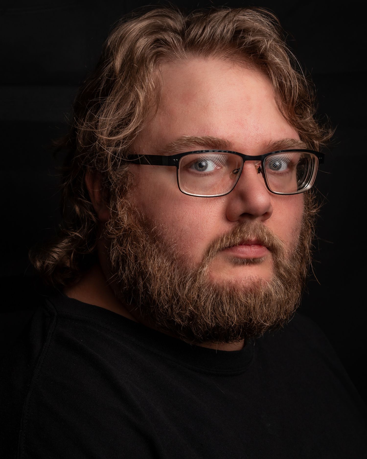 Bearded man with glasses and medium-length brown hair posing against a dark studio background.