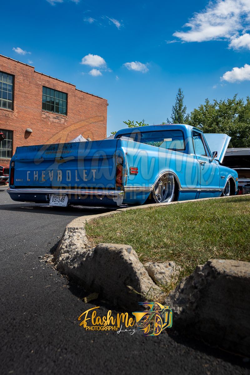 A classic blue Chevrolet pickup truck with a lowered stance parked at an outdoor car show on a sunny day.