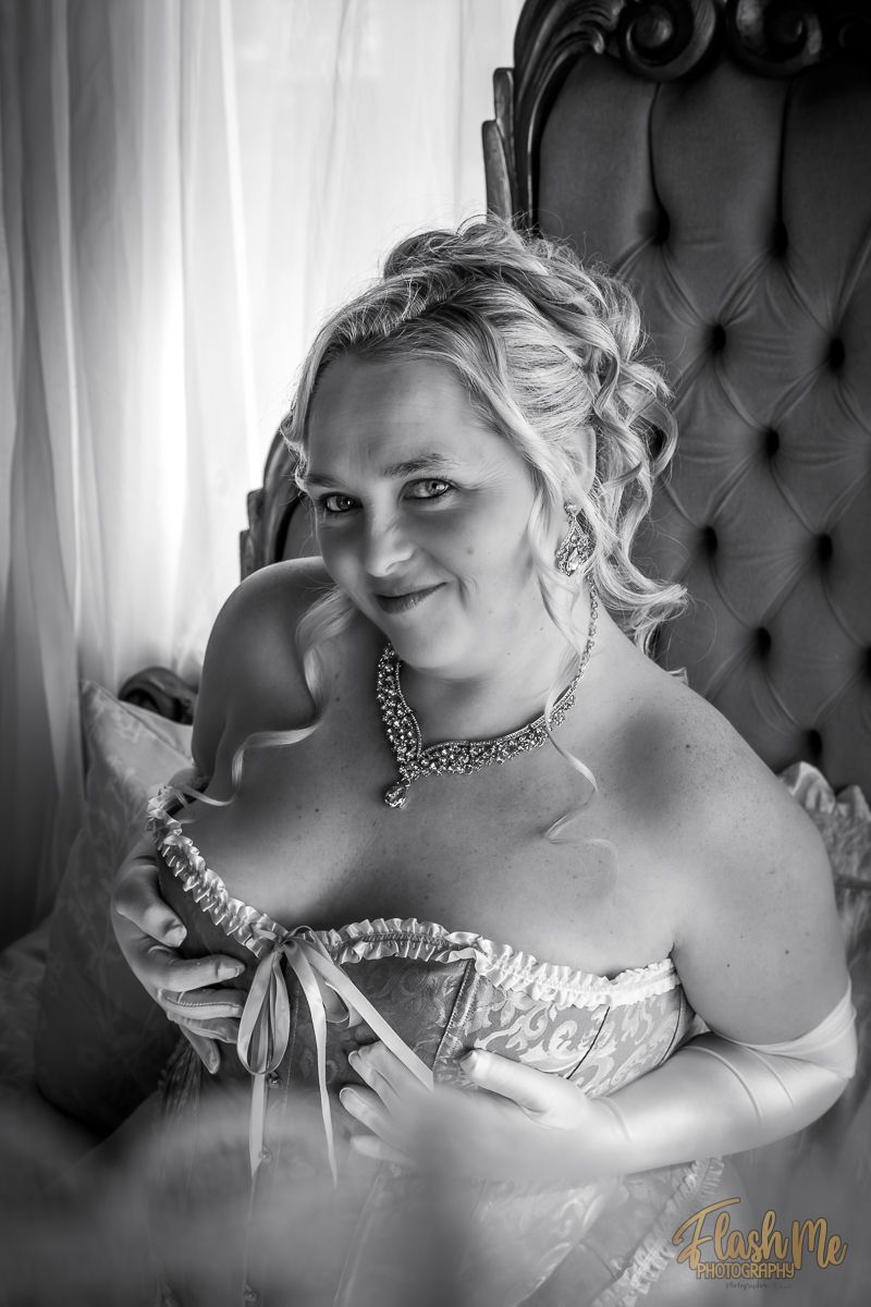 Black and white boudoir photo of a blonde woman in a corset sitting in a tufted chair holding a masquerade mask.