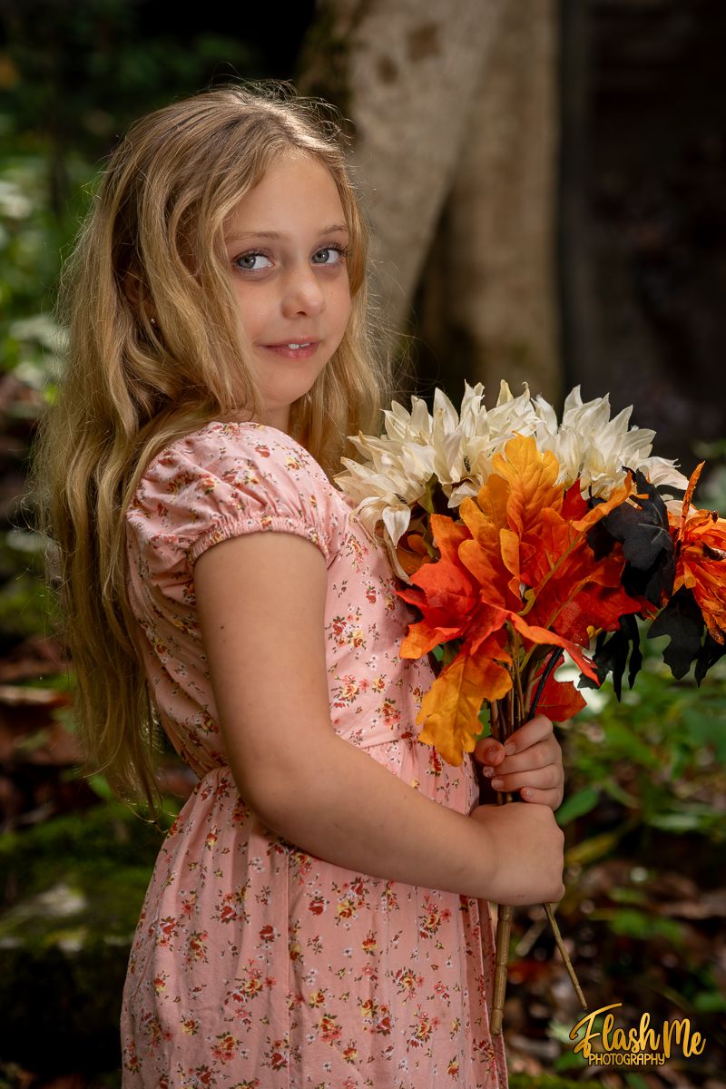 Young girl in pink floral dress holding a bouquet of colorful autumn flowers in a forest setting.