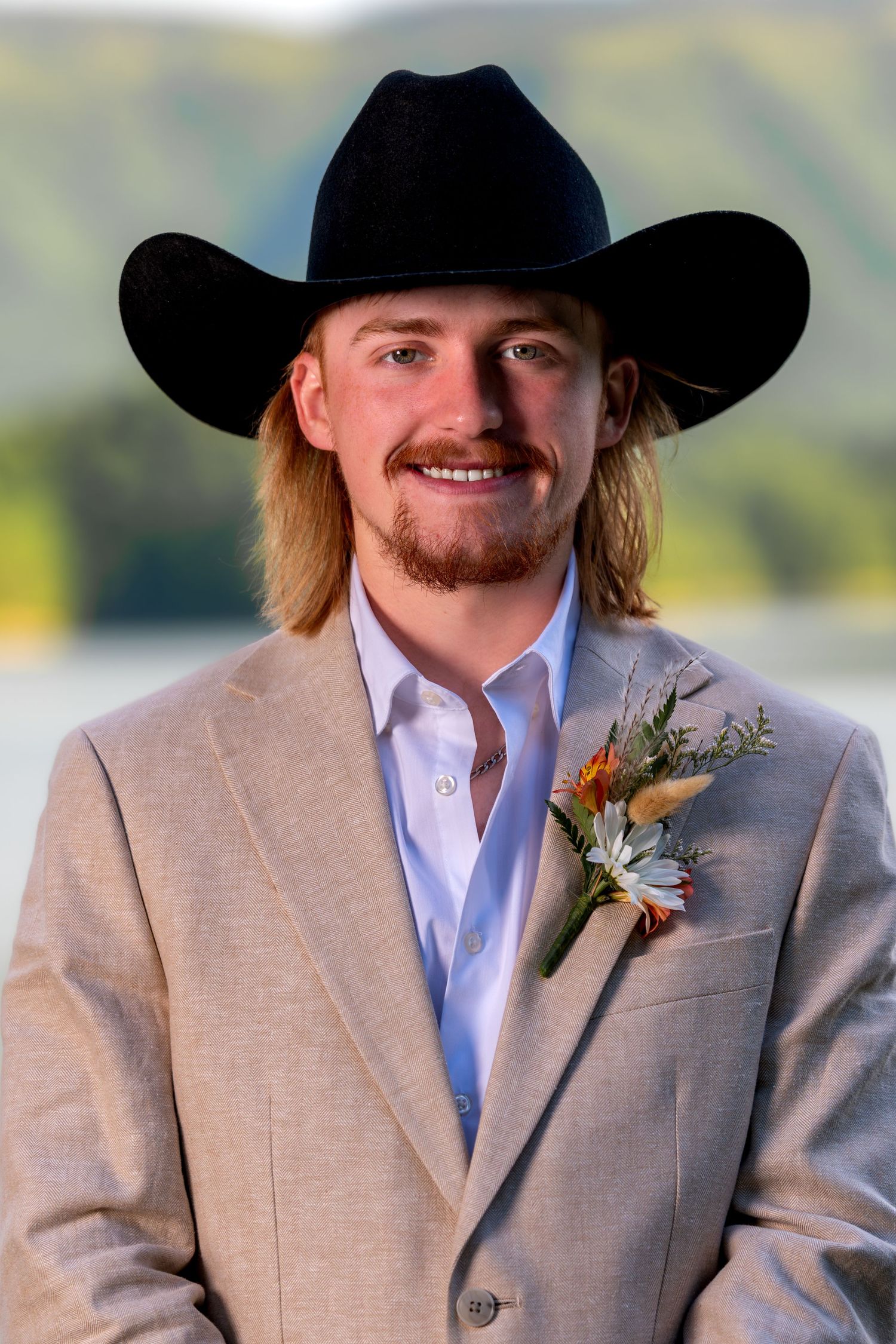Young man in tan suit and black cowboy hat with boutonniere smiling outdoors by a lake.