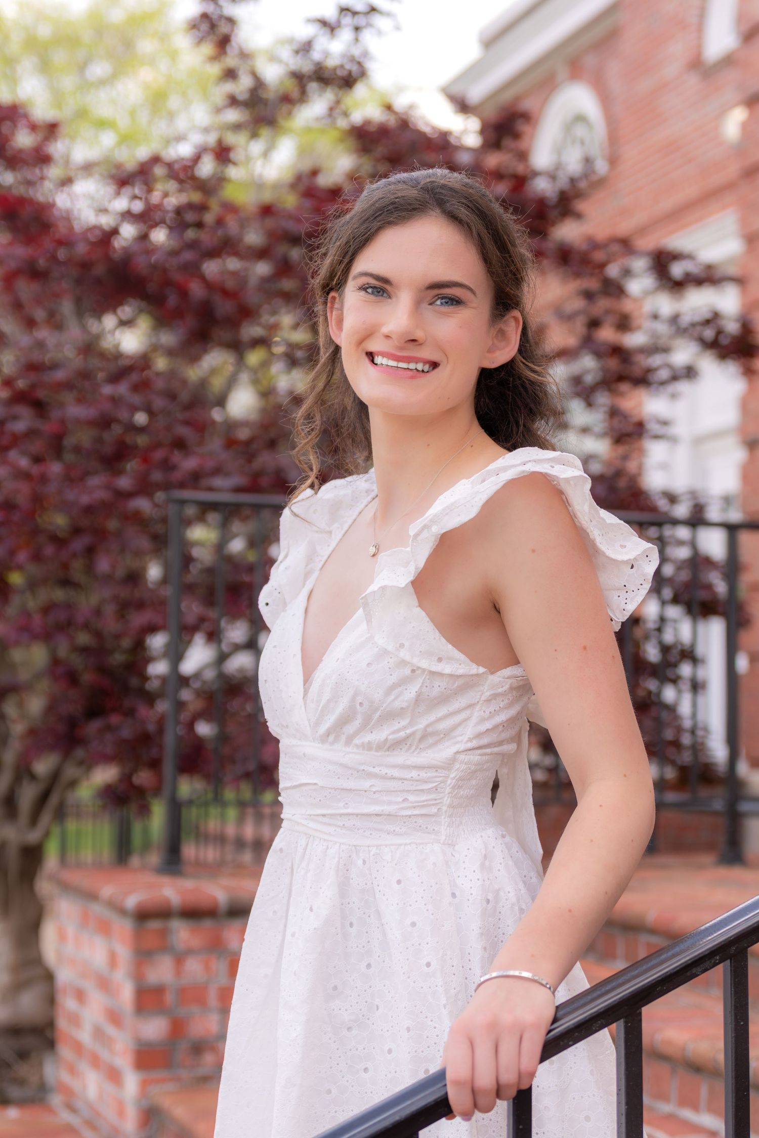 Smiling young woman in a white ruffled dress standing by a railing with red maple trees in background.