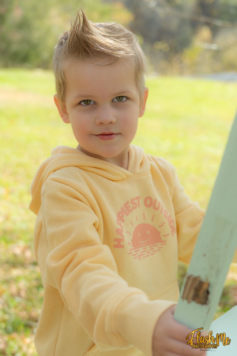 A young blonde boy in a yellow hoodie poses outdoors in a sunny autumn park setting.