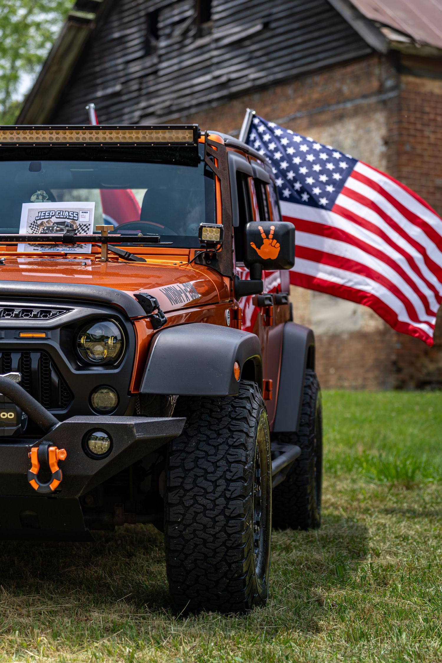 Orange Jeep Wrangler with American flag and off-road accessories parked on grass near a barn.