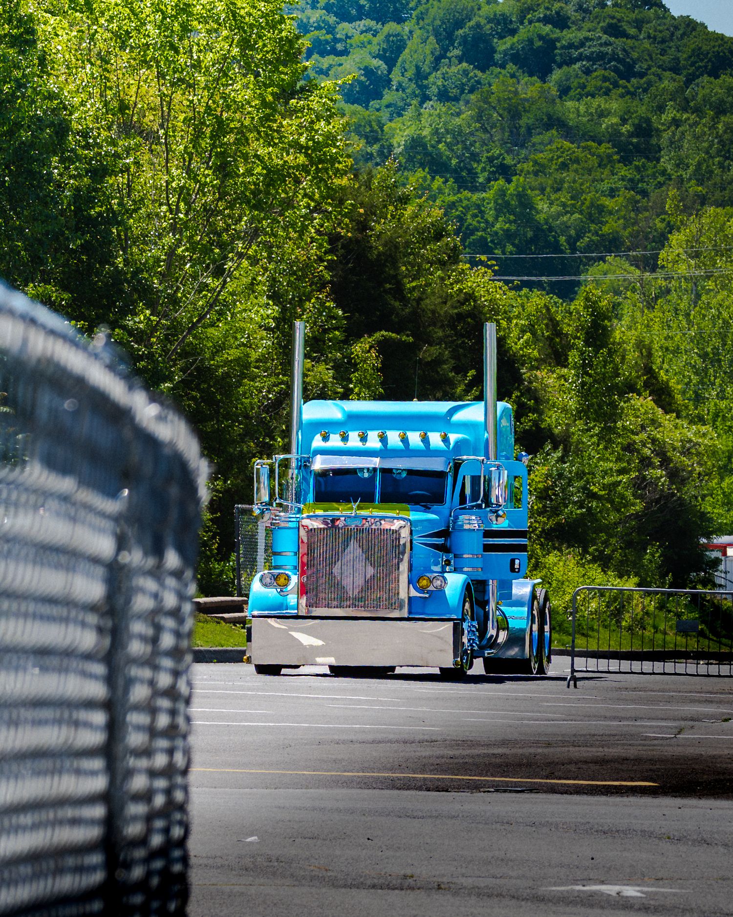 Bright blue Peterbilt semi truck driving on a road surrounded by green trees.