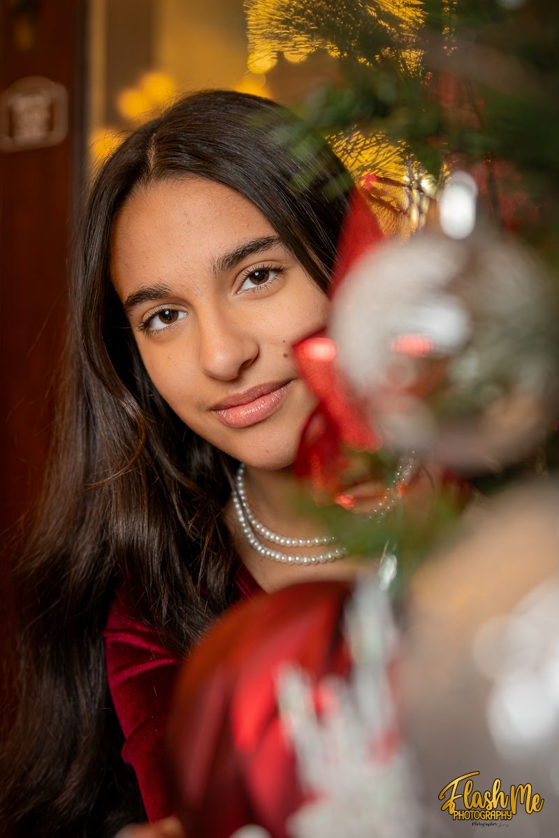 Young teenage girl in red velvet dress smiles beside a decorated Christmas tree with ornaments.