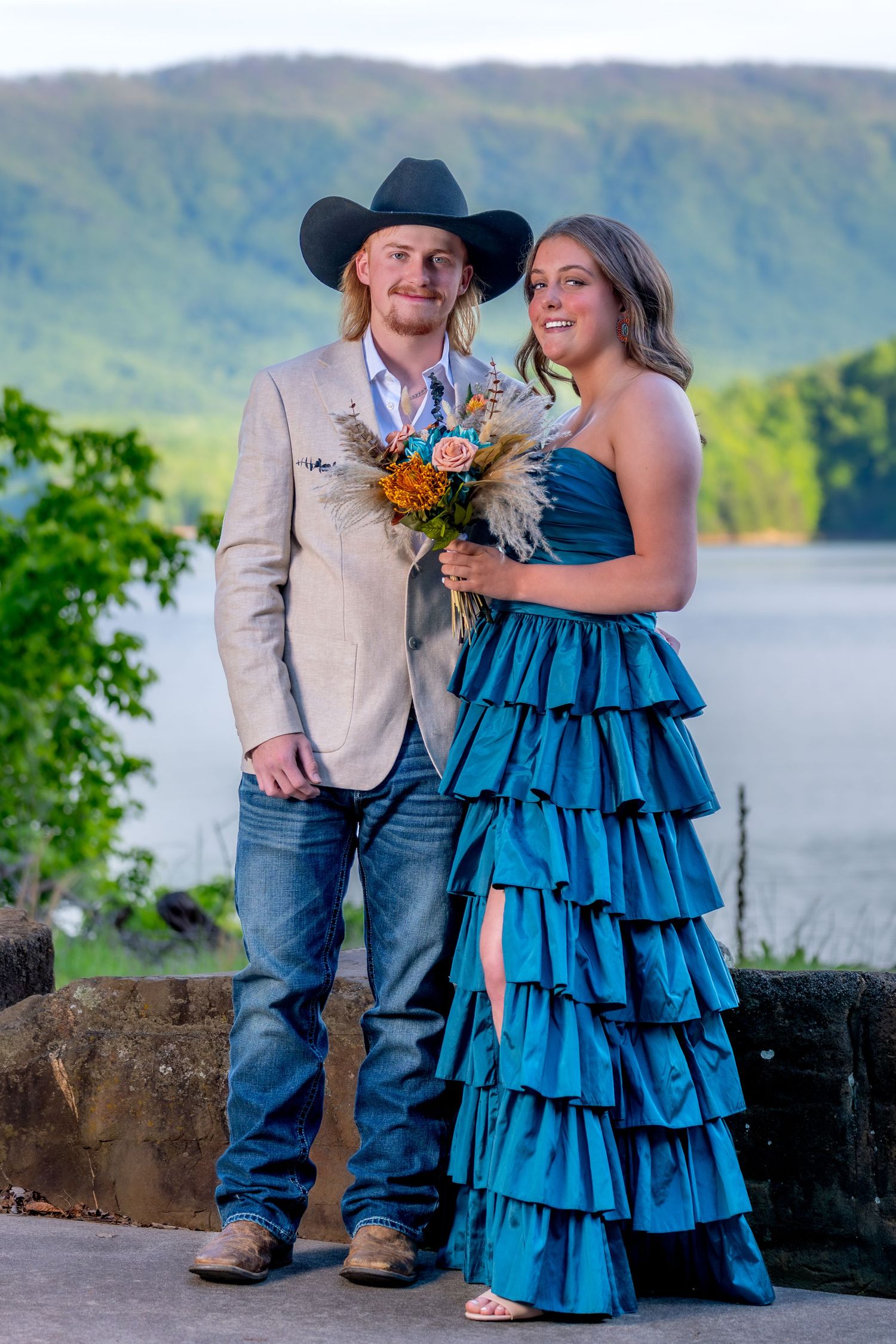 Couple posing outdoors by a lake, man in cowboy hat and tan suit, woman in blue ruffled gown.