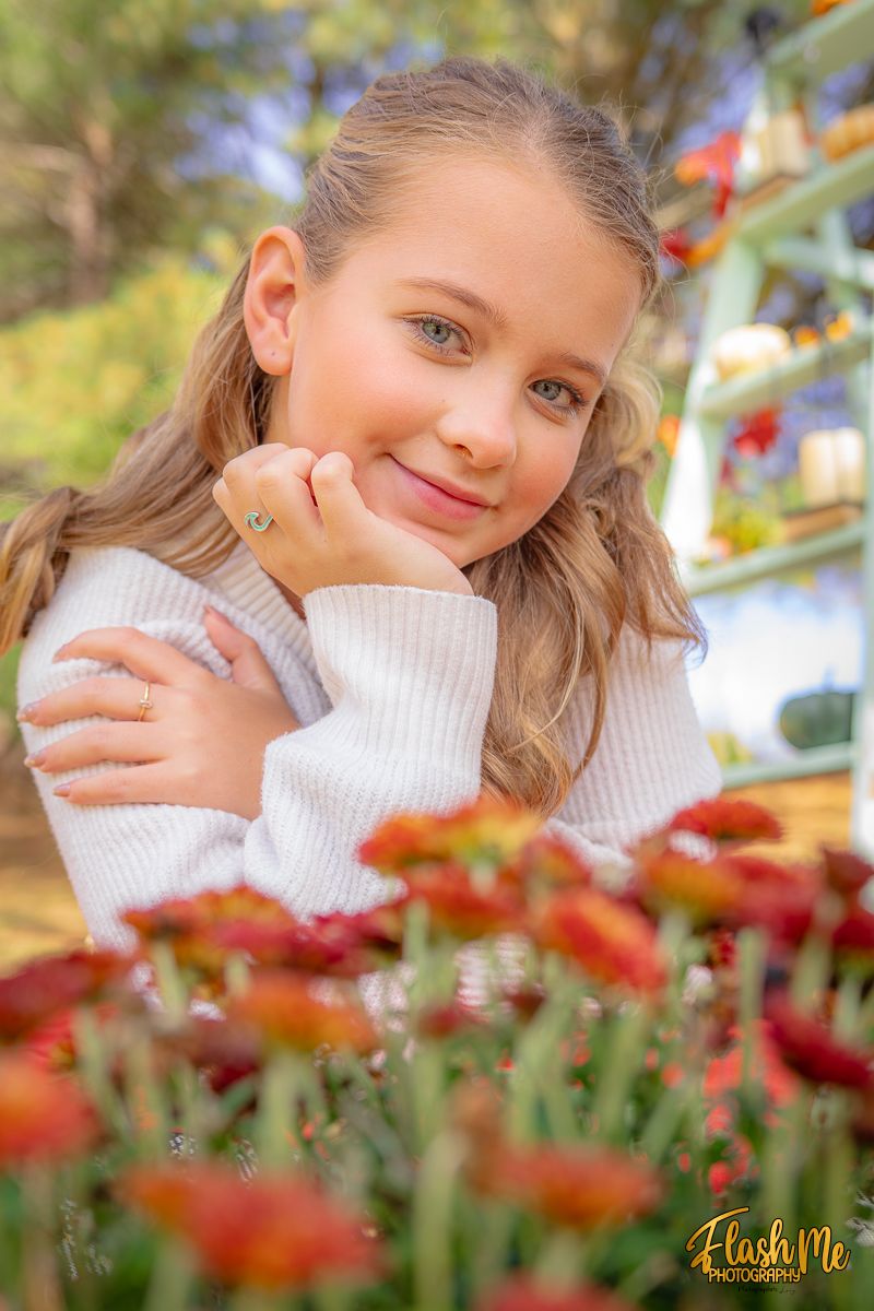 A smiling young girl in a white sweater rests her chin on her hand near colorful fall flowers.