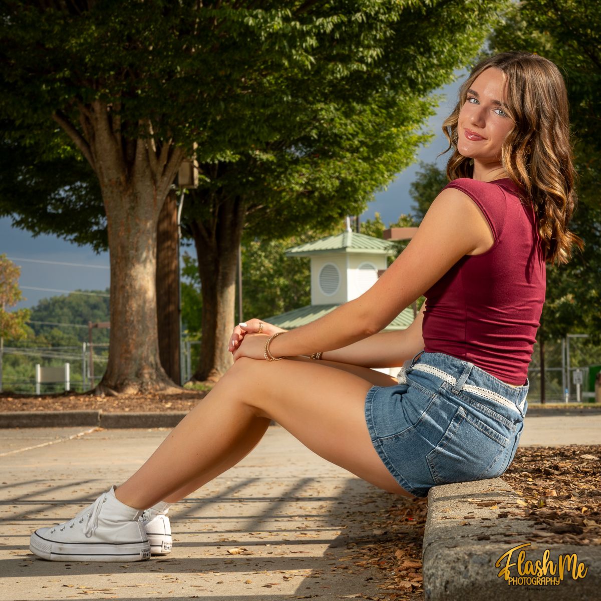 Young woman in maroon top and denim shorts sitting outdoors on a sunny day wearing white Converse sneakers.