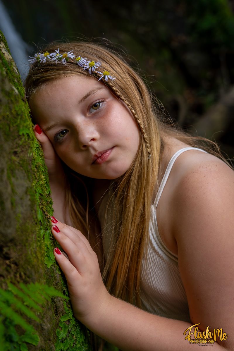 Young girl wearing daisy headband leaning against moss-covered rock near a waterfall in the forest.