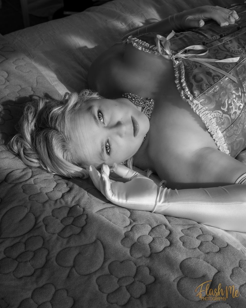 Black and white photo of a blonde woman in lace lingerie and satin gloves lying on a bed.