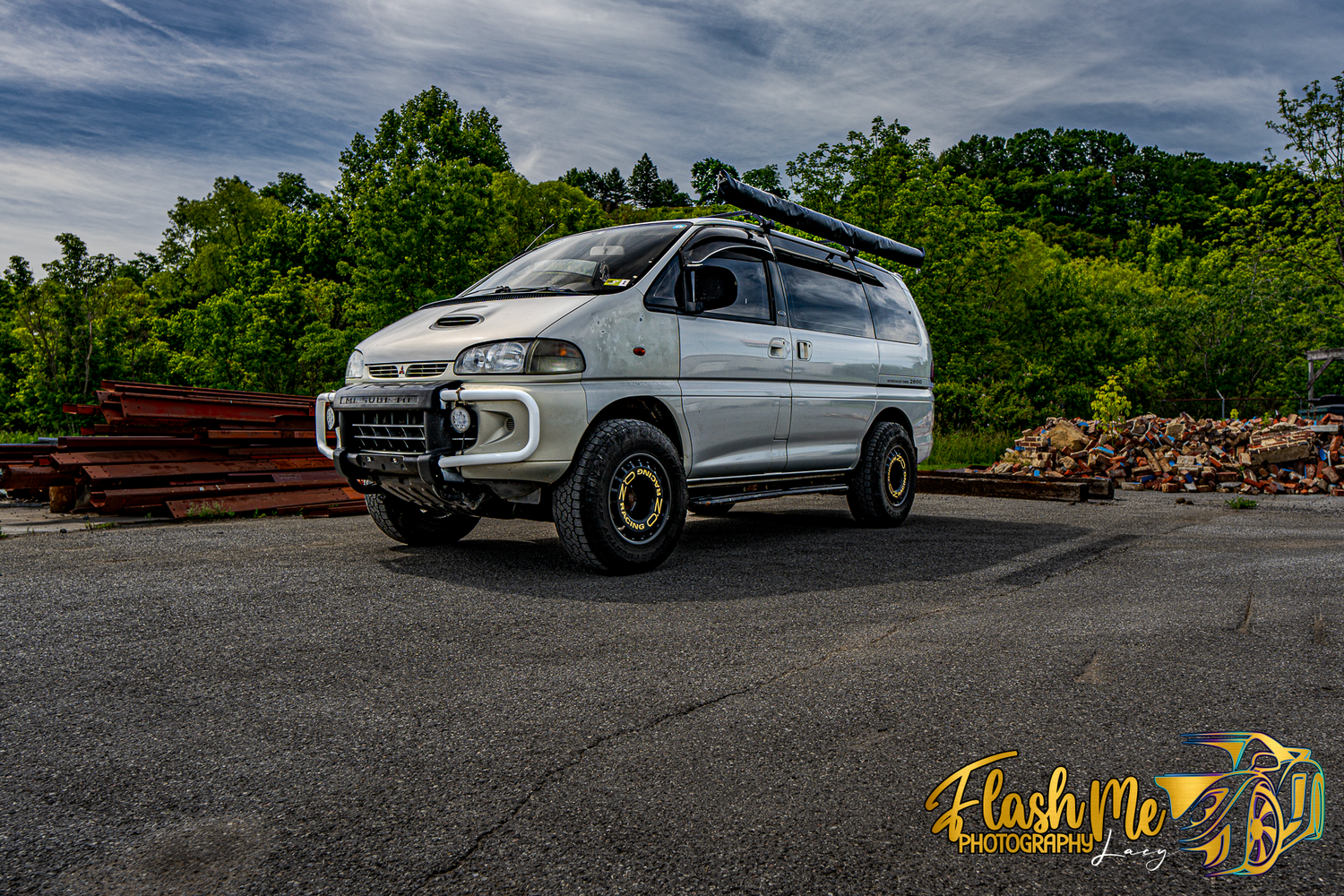 White Mitsubishi Delica off-road van parked on an empty lot surrounded by lush green trees.
