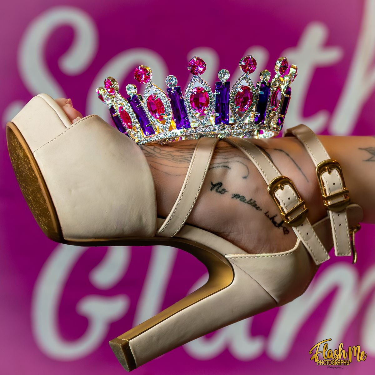 Bejeweled crown resting on a beige high heel shoe with ankle straps and a tattoo visible on the foot.