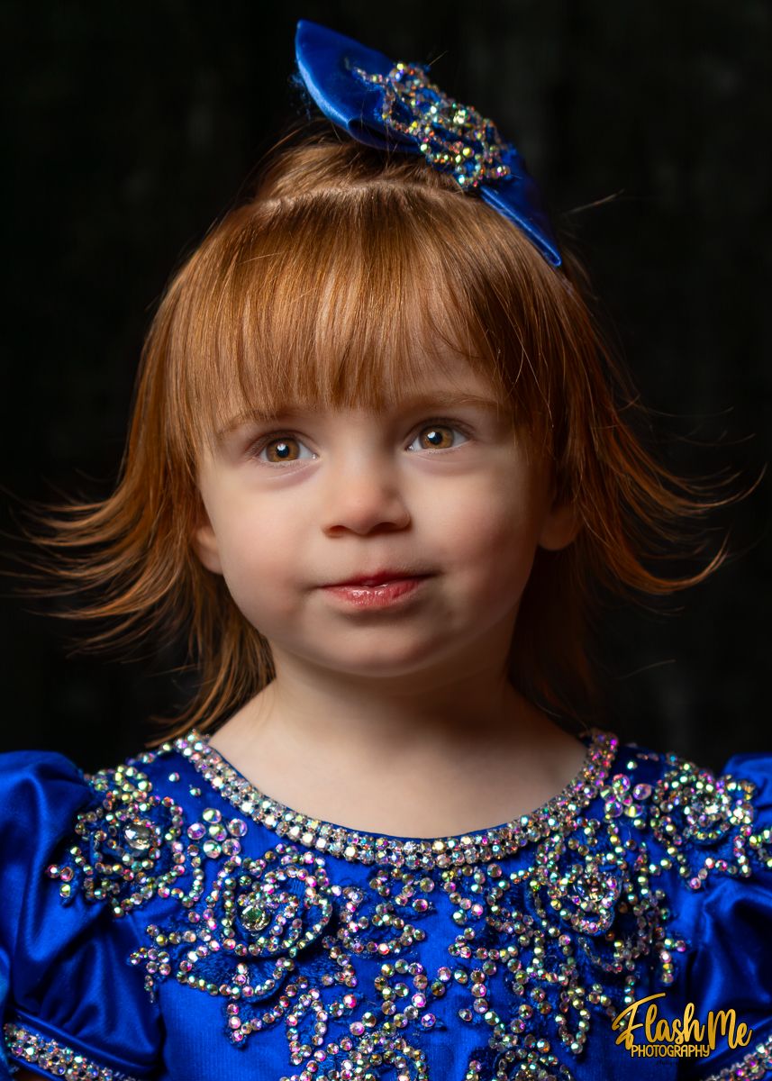 Young redhead girl in blue rhinestone pageant dress with bow headpiece in studio portrait.