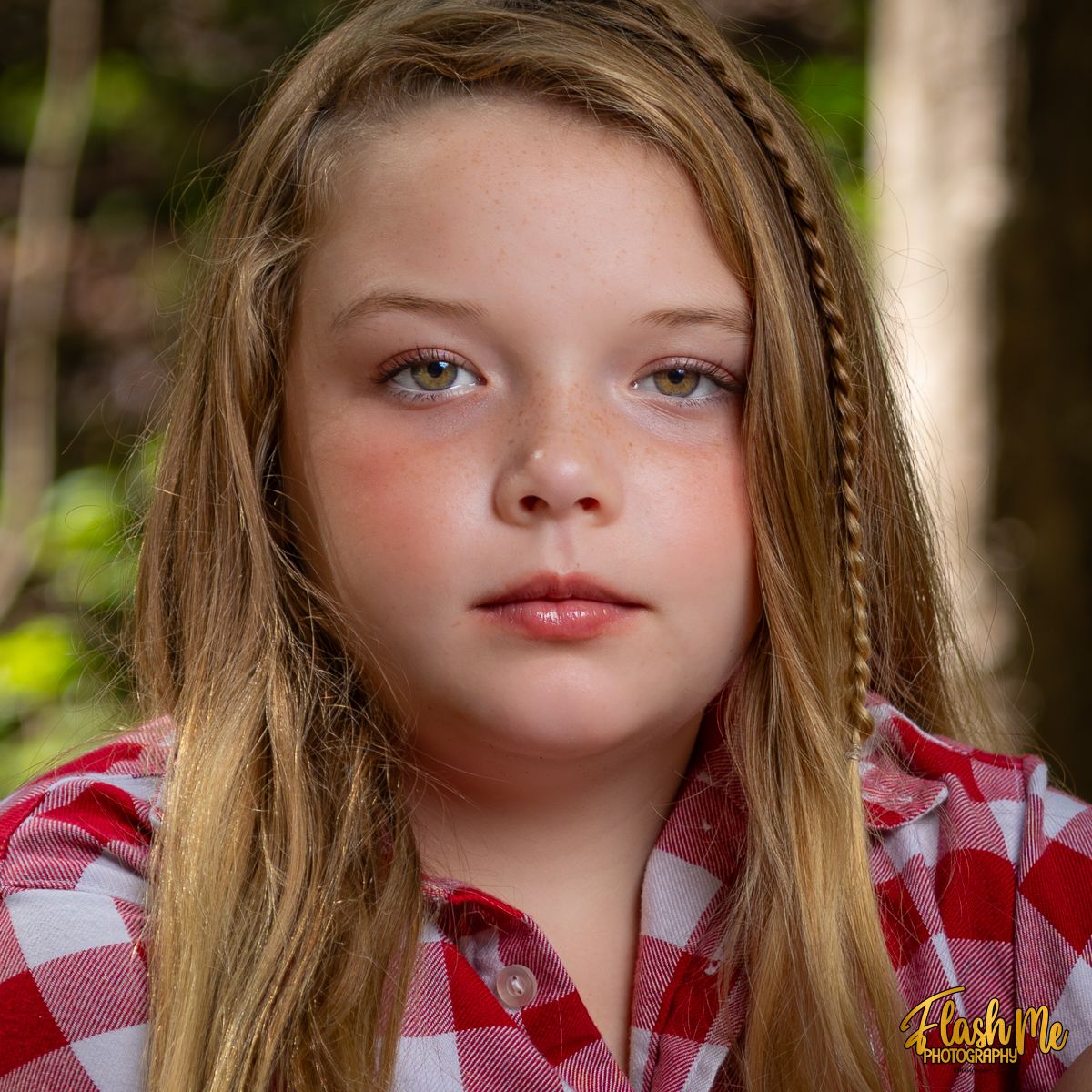 Close-up portrait of a young girl with long blonde hair and braids wearing a red plaid shirt outdoors.