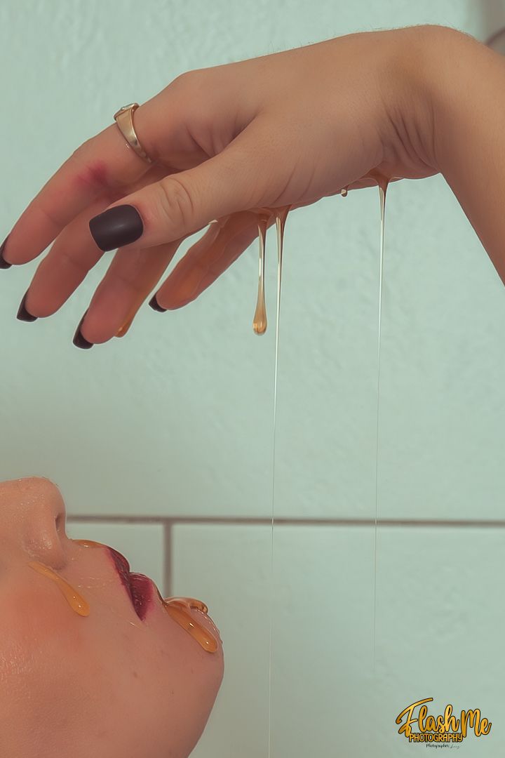 Honey dripping from a hand with black nails and gold ring onto a persons face below.