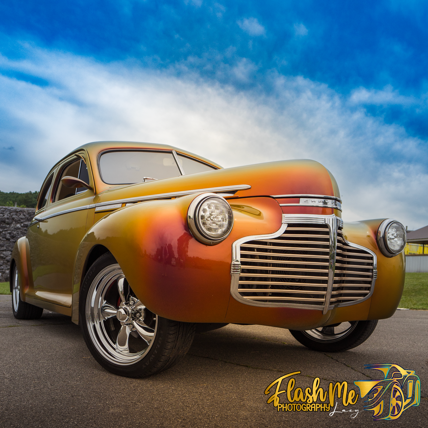 Golden orange custom classic car with chrome grille under a dramatic blue sky.