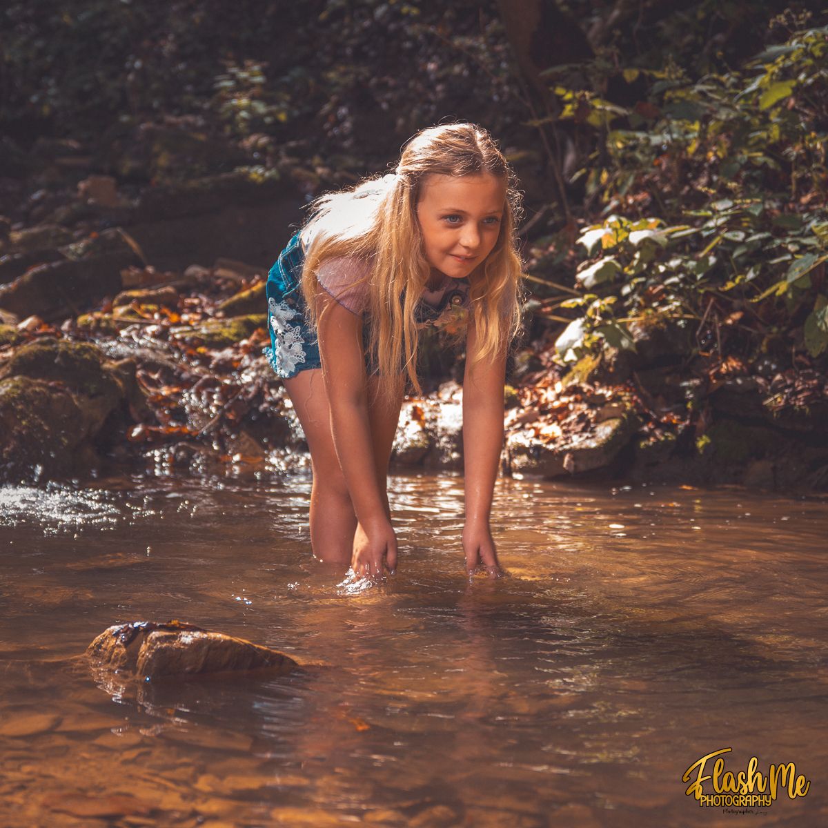 Young blonde girl smiling while playing in a shallow sunlit forest stream on a sunny day.