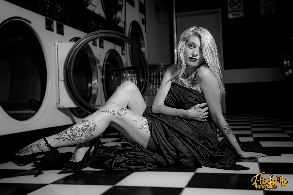 Black and white photo of a tattooed blonde woman in a black dress posing in a laundromat.