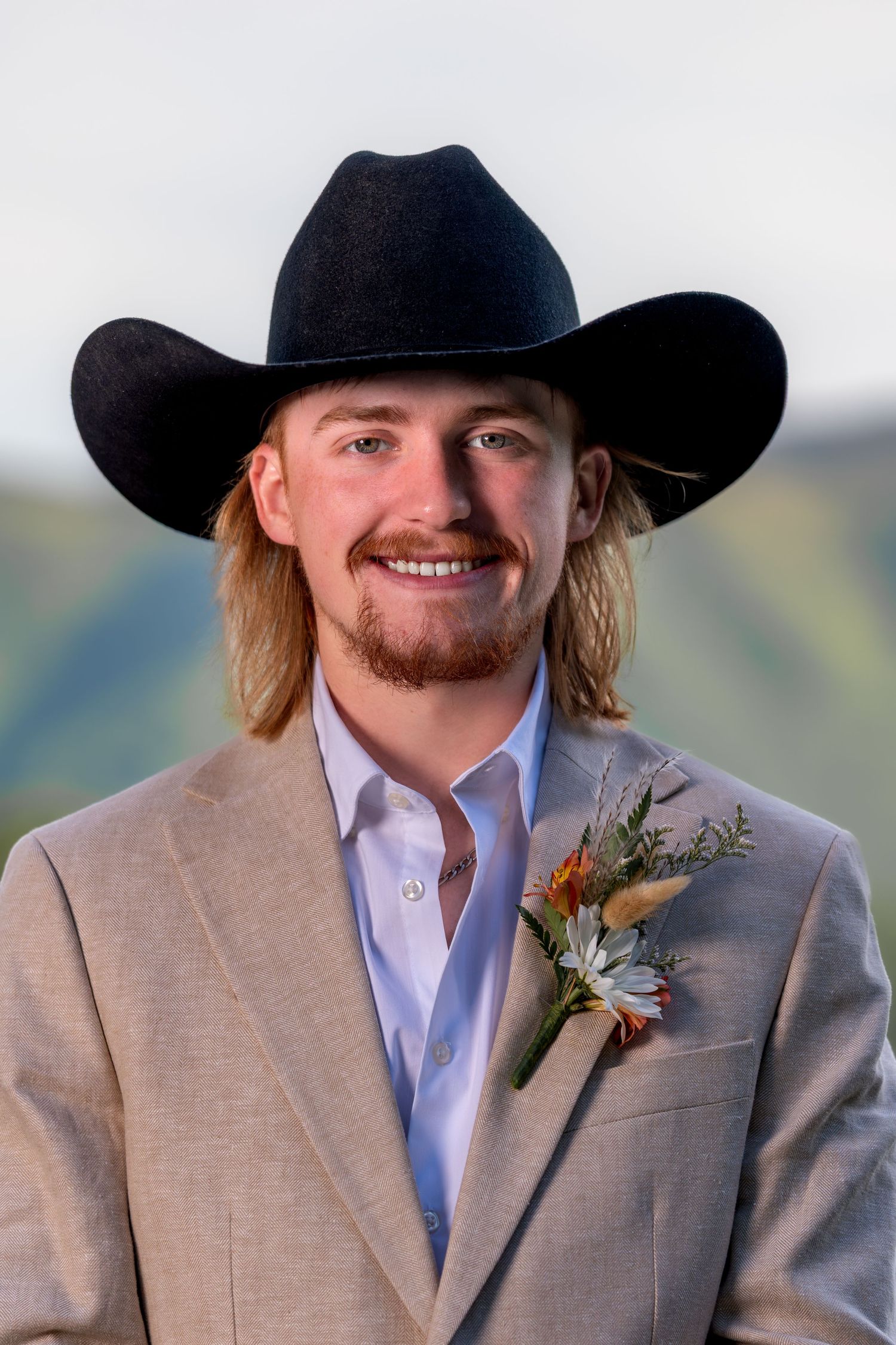 Smiling man in black cowboy hat and tan suit with floral boutonniere at outdoor event.