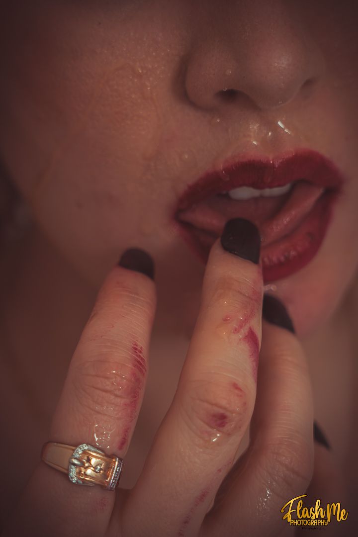 Close-up artistic photo of a woman with red lips and dark nails touching her face with ringed fingers.