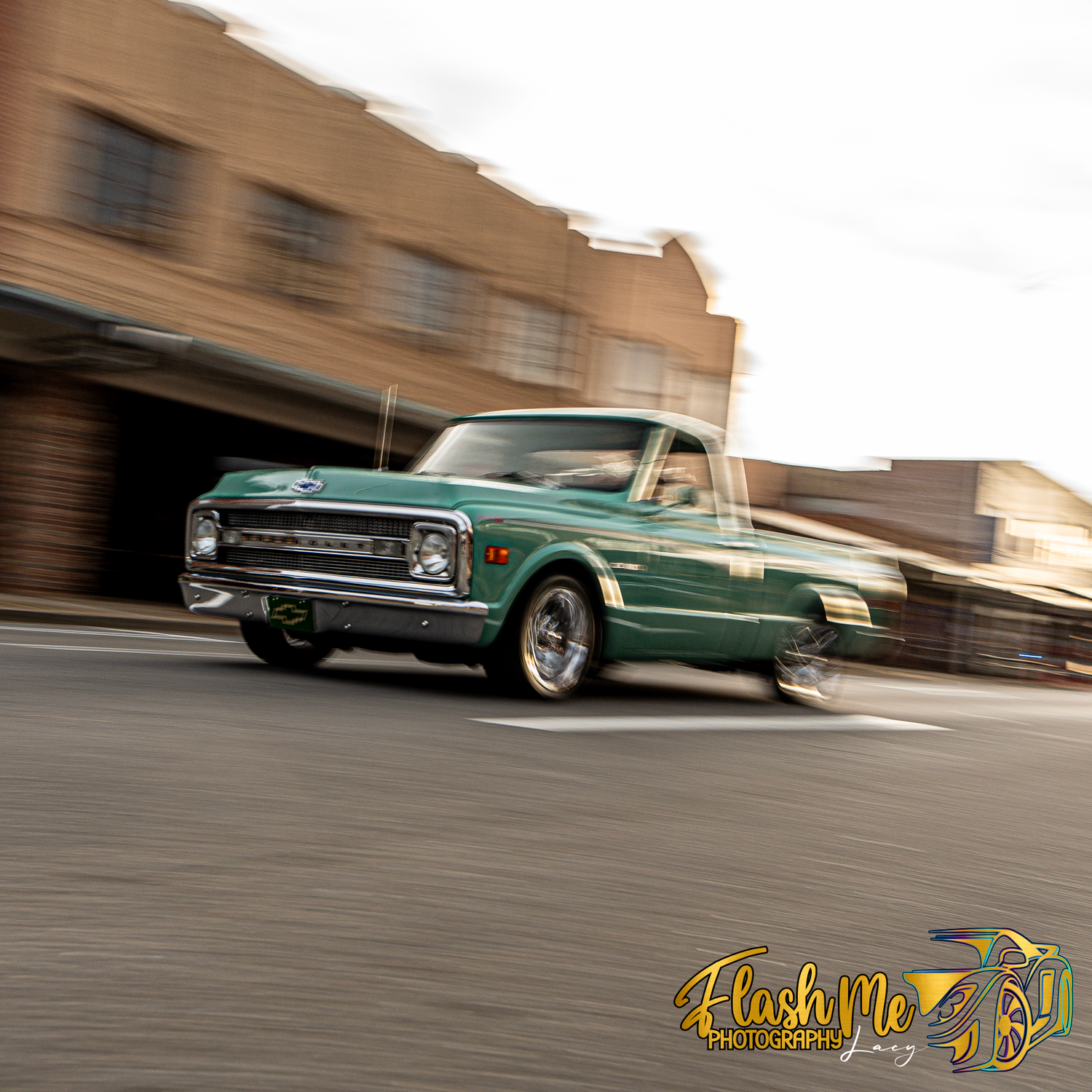Green classic Chevrolet C10 pickup truck driving through a downtown street with motion blur.