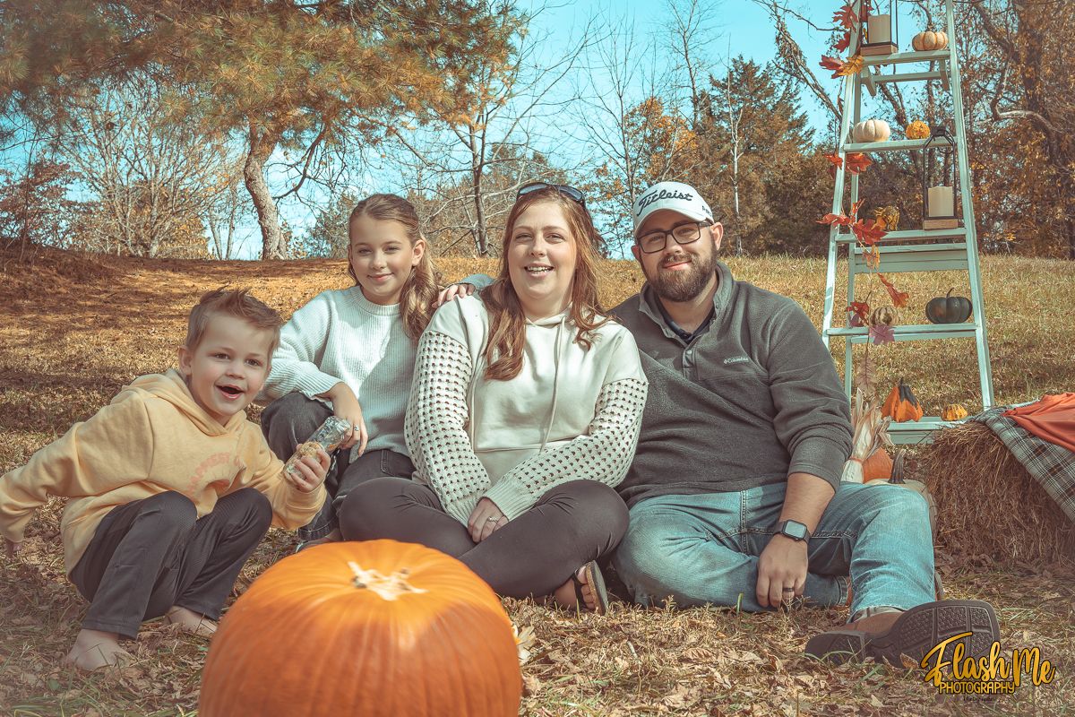 A happy family of four sits together outdoors surrounded by pumpkins during a fall photo session.