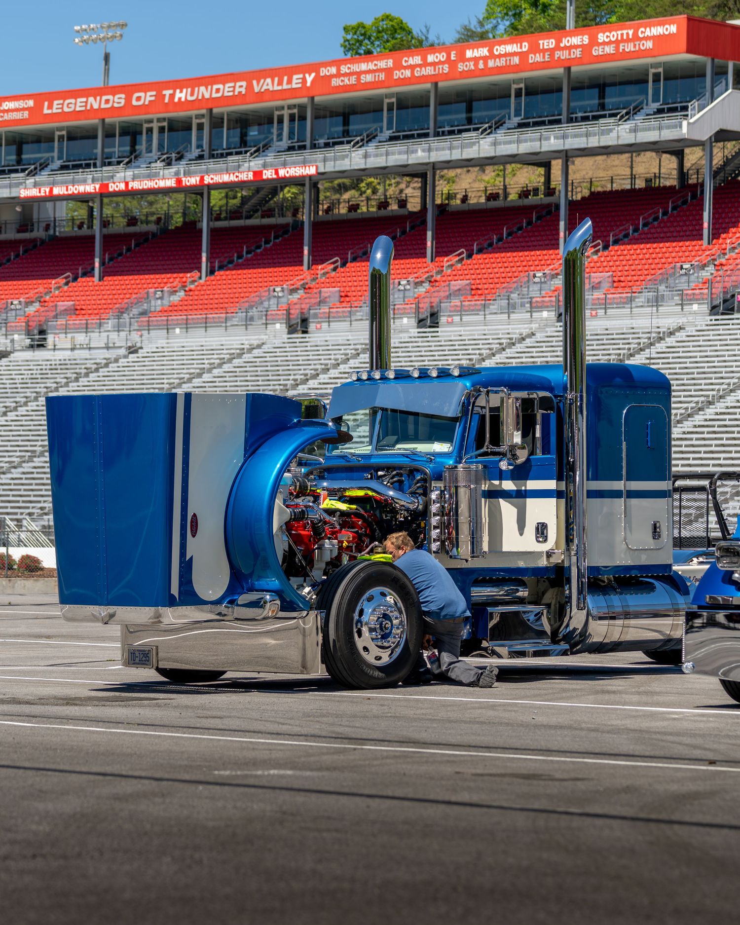 Blue custom show semi truck with open hood on display at a NASCAR speedway venue.