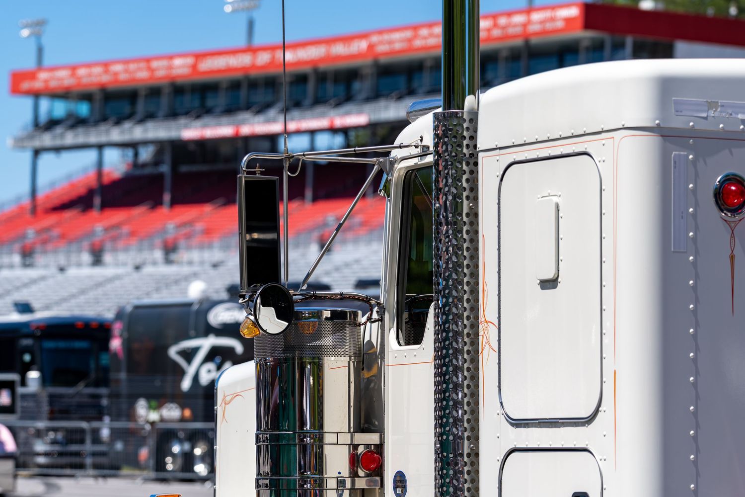 A white semi-truck cab parked near a NASCAR racing stadium on a bright sunny day.