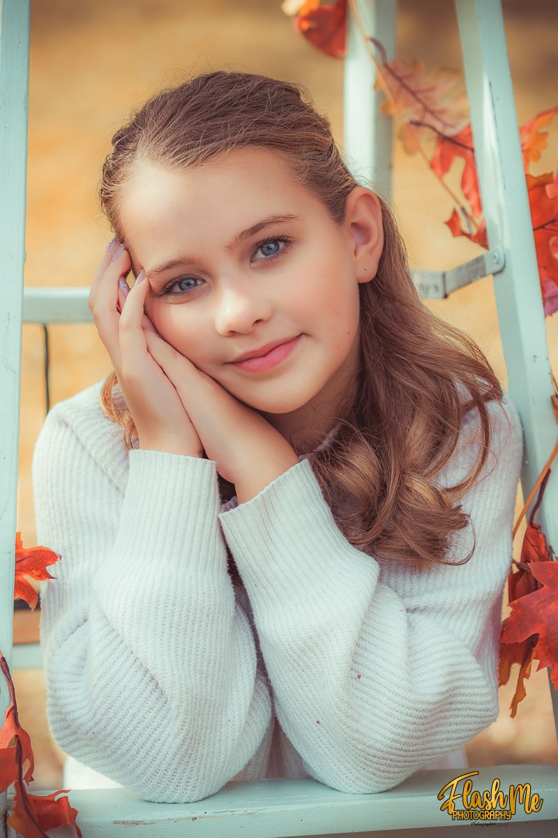 A pretty young girl with blue eyes rests her face in her hands surrounded by colorful autumn leaves.
