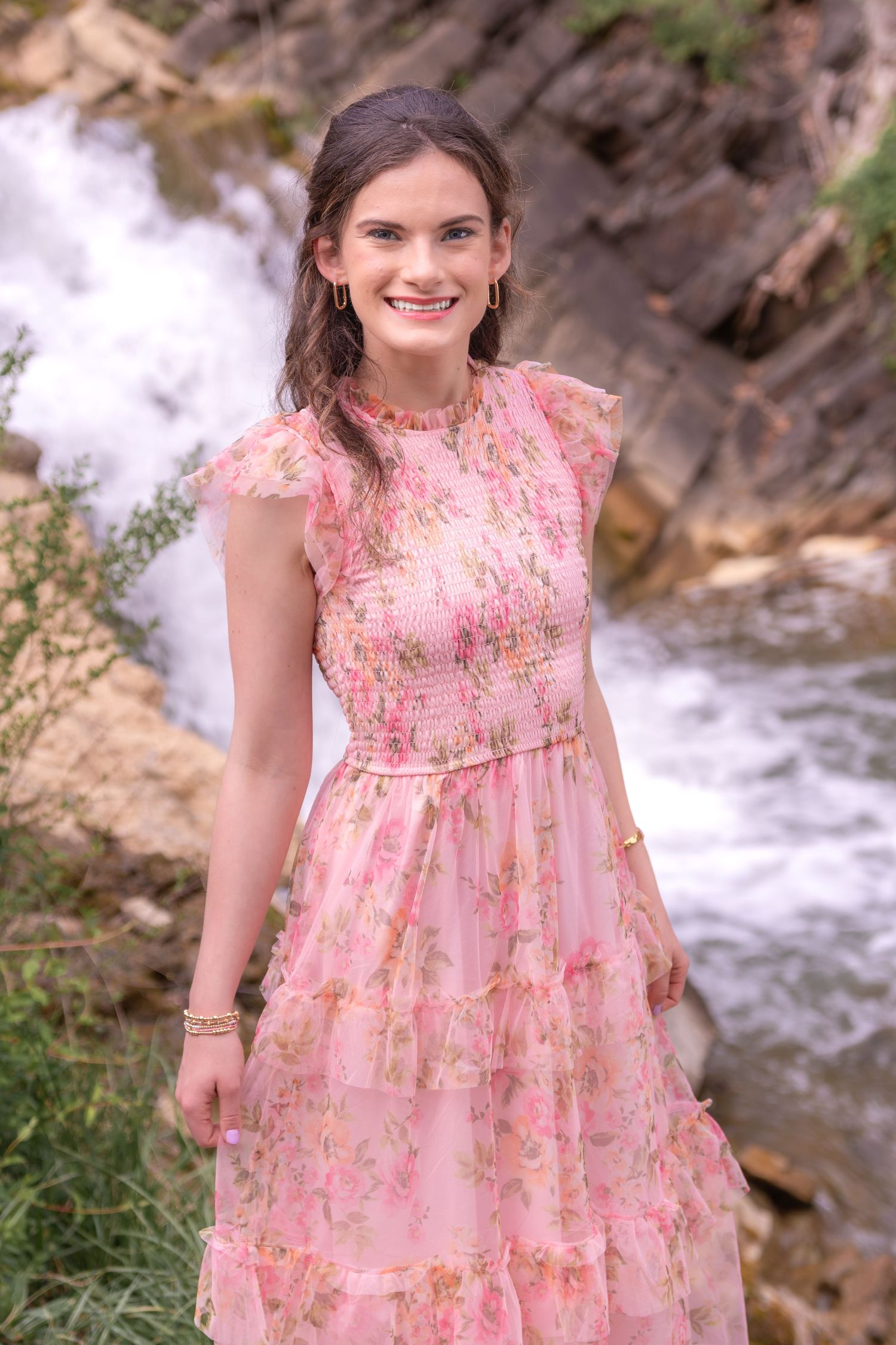 Smiling young woman in a pink floral tiered dress standing near a waterfall surrounded by rocks and greenery.