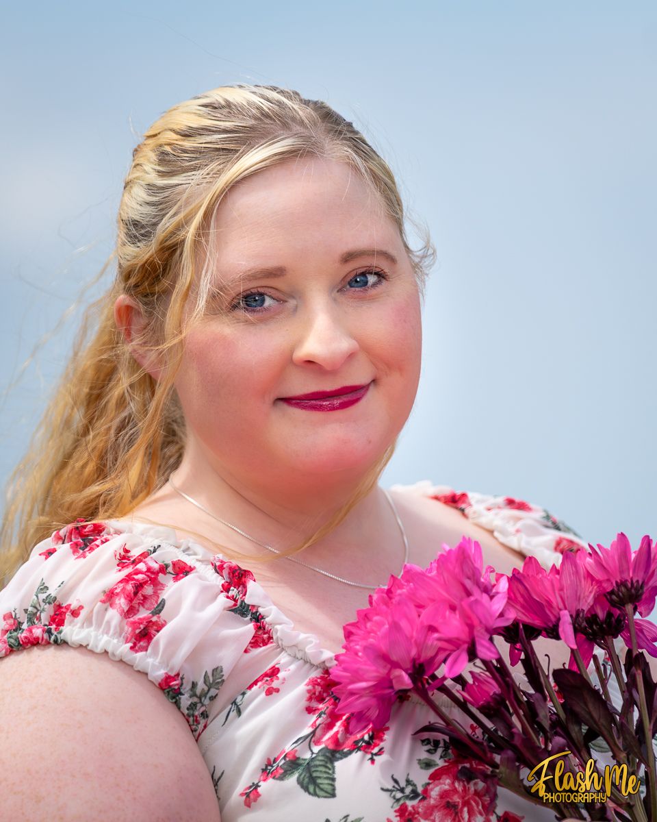 Smiling blonde woman with blue eyes holding pink flowers wearing a floral dress against a light blue sky.