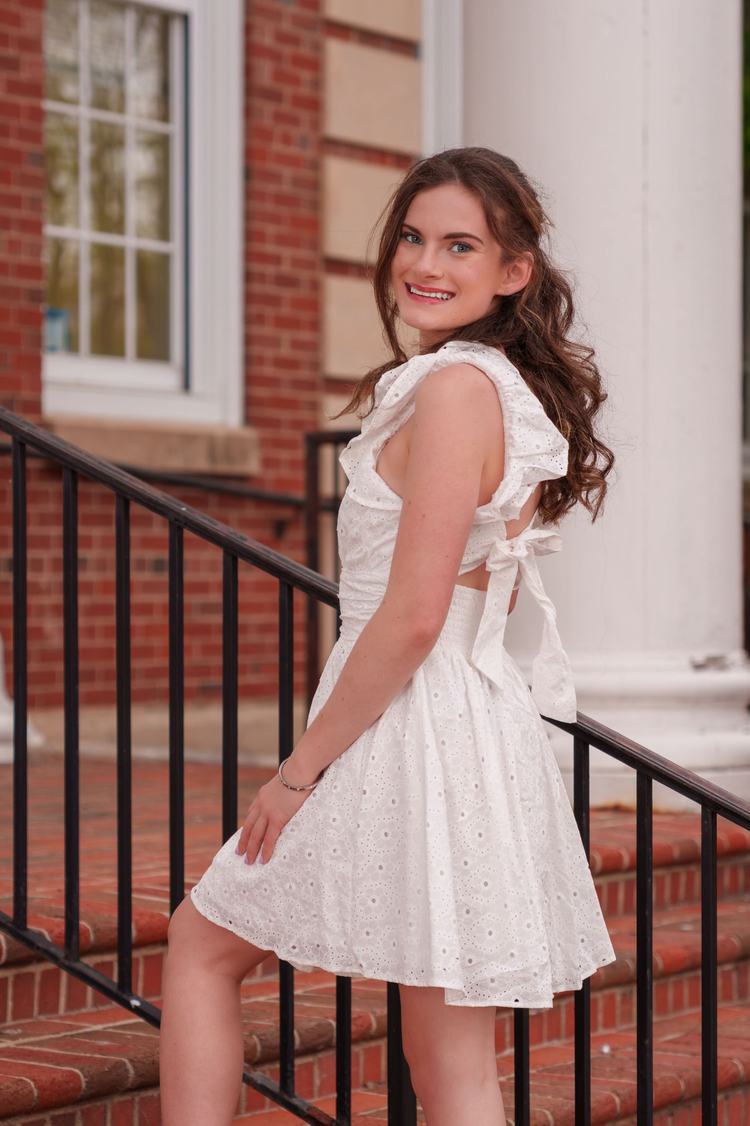 Smiling young woman in a white eyelet dress posing on brick steps outside a classic building.