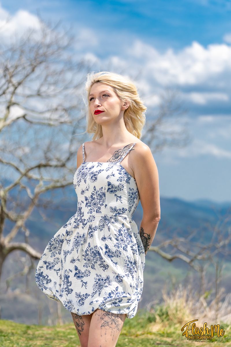 Blonde woman in floral sundress standing on mountain overlook under dramatic cloudy sky.