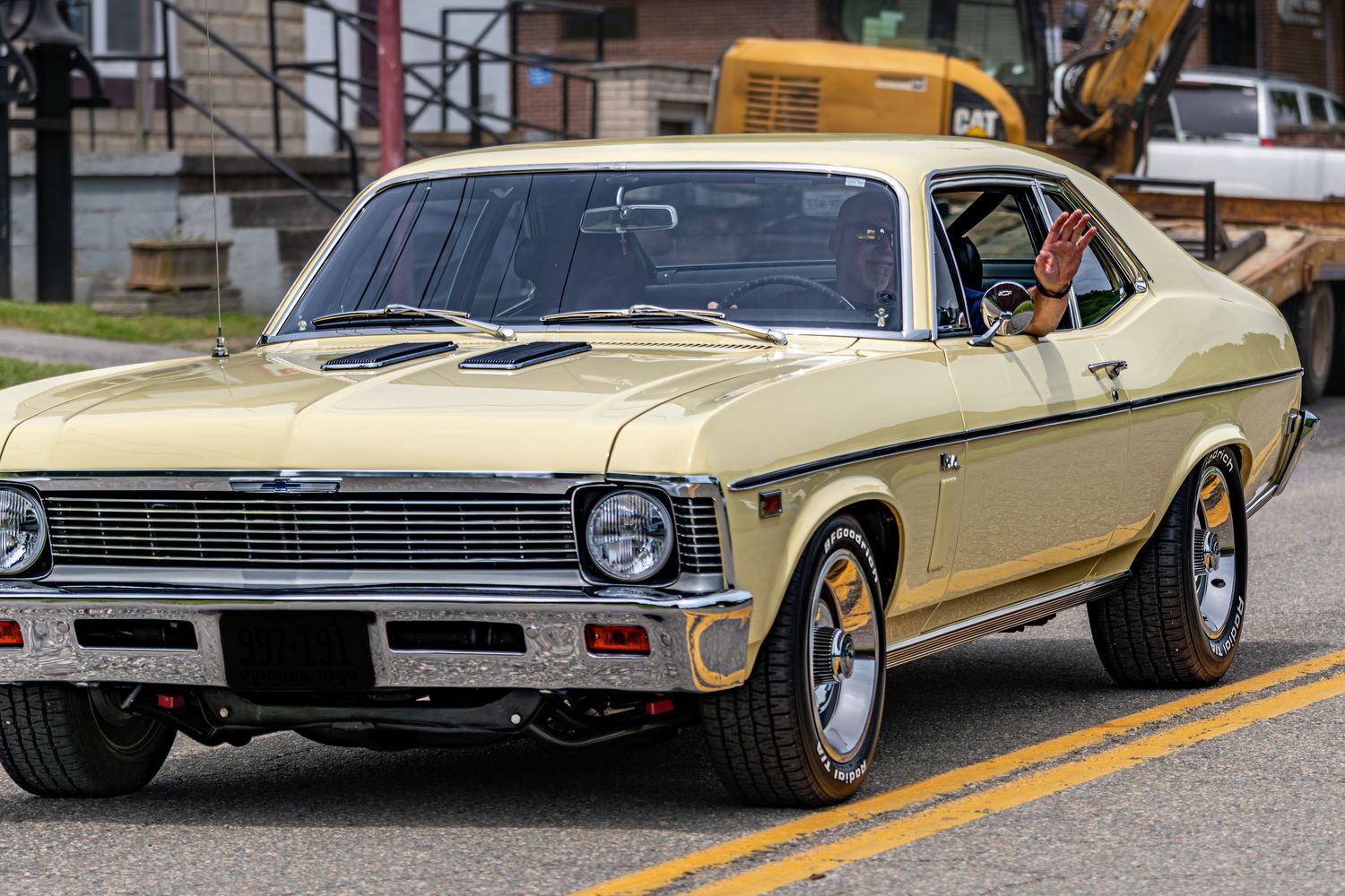 Classic yellow Chevrolet Nova muscle car driving on a street with chrome wheels and hood scoop.