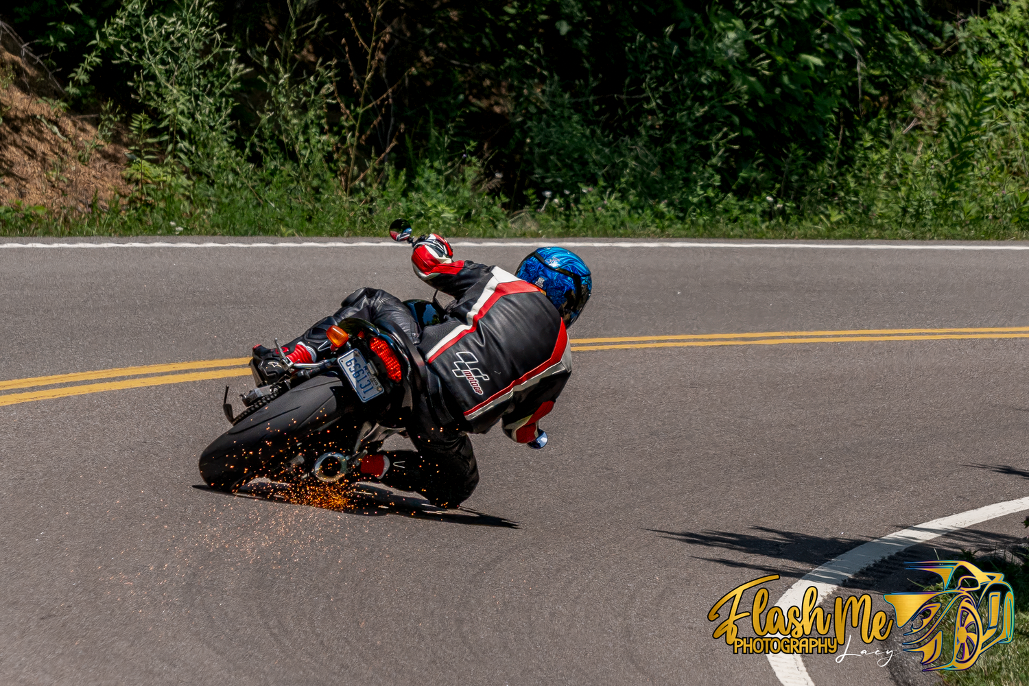 Motorcycle racer leaning into a sharp curve on a tree-lined road during a race event.