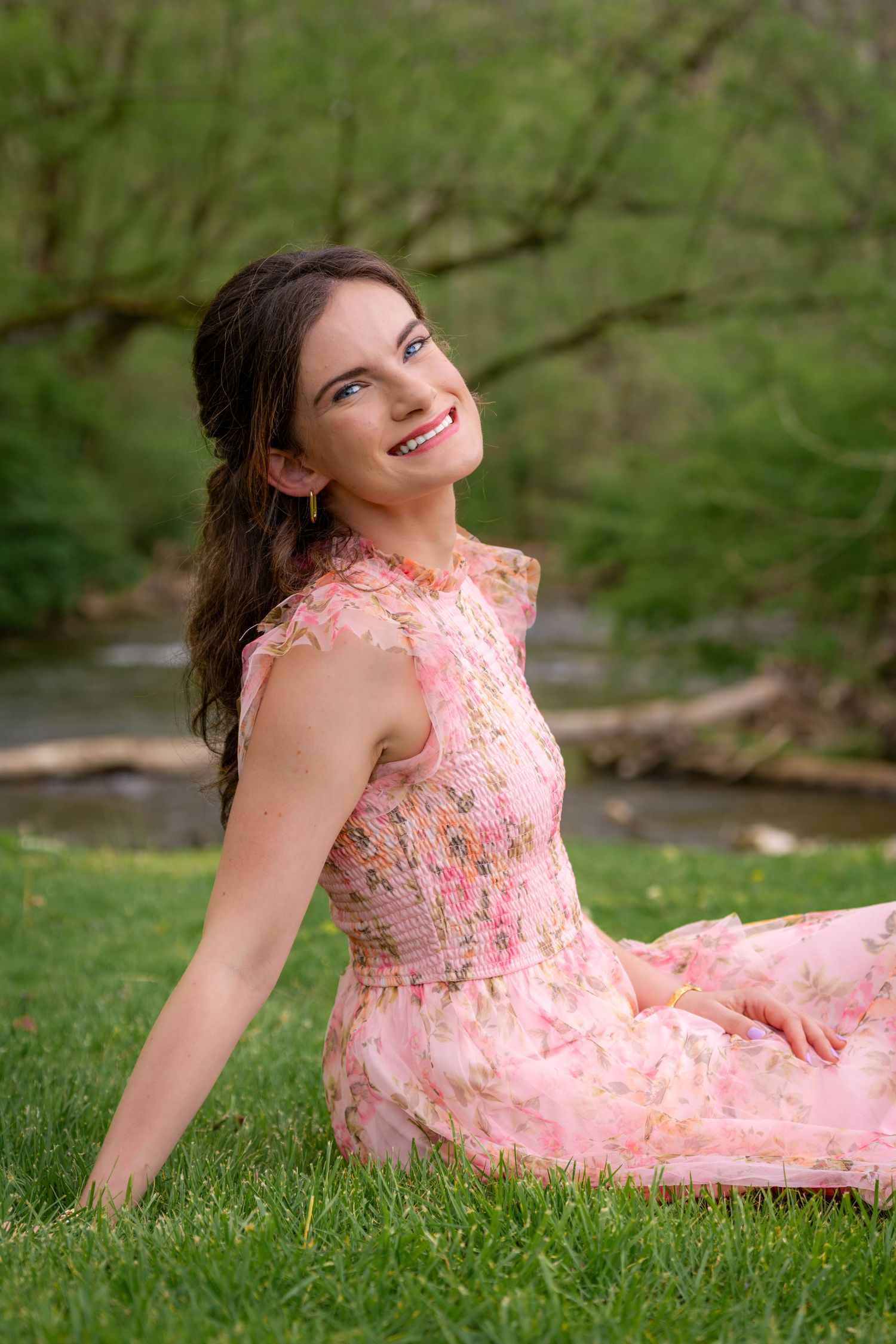 Young woman in a pink floral dress sitting on green grass near a stream smiling upward joyfully.