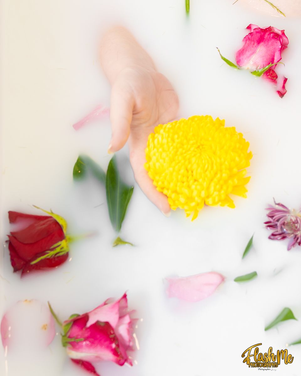 A hand holds a yellow chrysanthemum surrounded by colorful roses and petals floating in a milk bath.