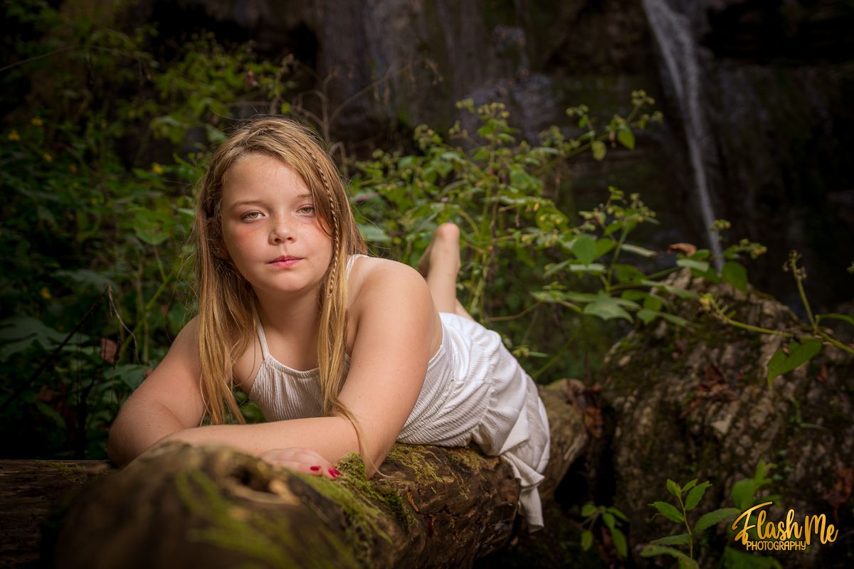 Young girl in white dress lying on a mossy log with a waterfall and lush greenery in background.
