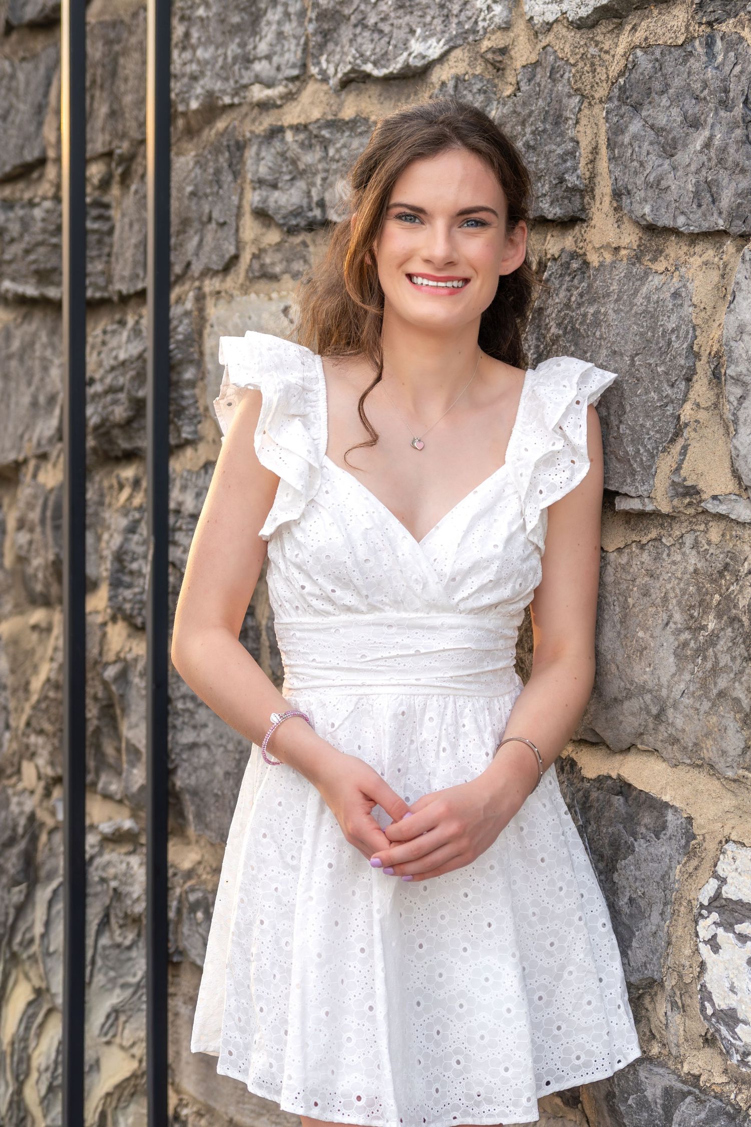 Smiling young woman in a white eyelet ruffle dress leaning against a stone wall outdoors.