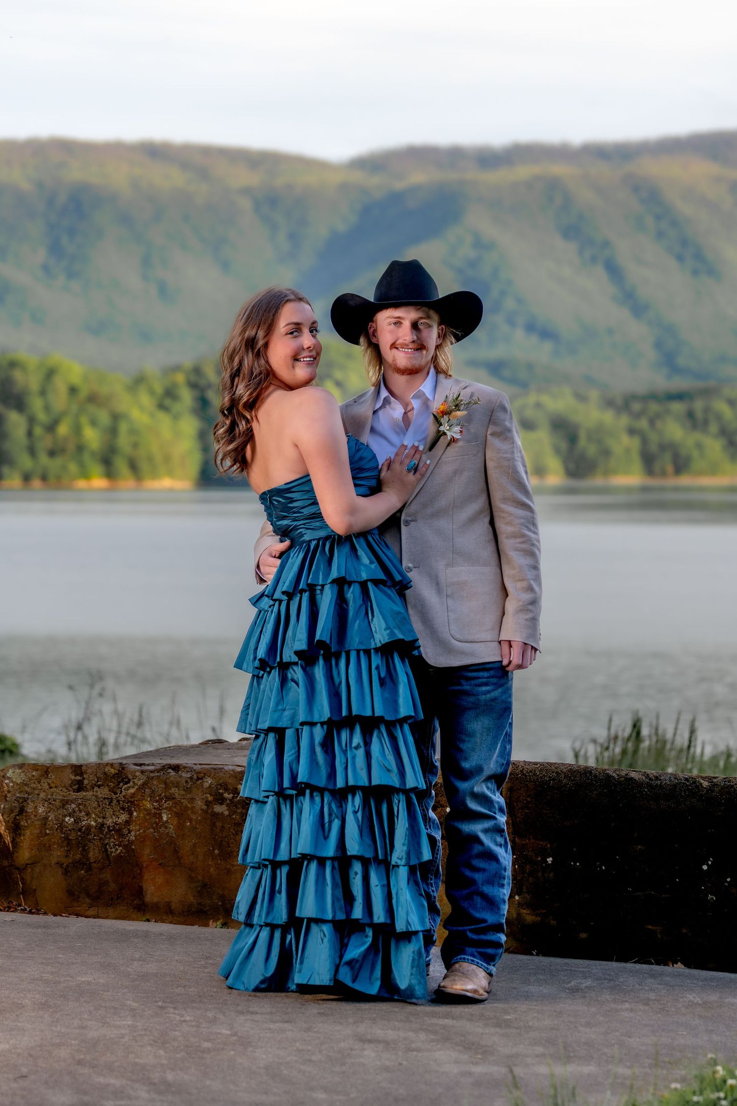 Prom couple posing by a lake with mountain views, wearing a teal ruffled gown and tan suit with cowboy hat.