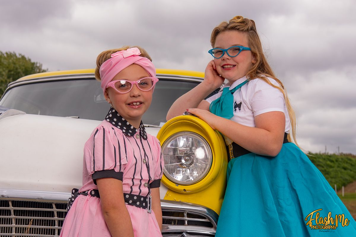 Two young girls in vintage 1950s-style outfits posing in front of a classic white car at an outdoor event.