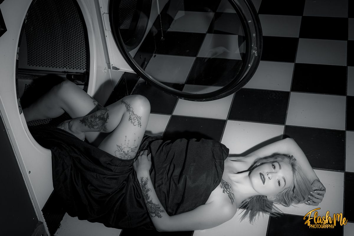 Black and white artistic photo of a tattooed woman in a black dress lying near a laundromat dryer.