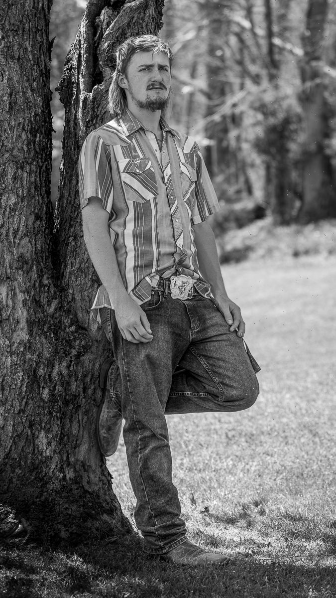Black and white portrait of a man leaning against a tree in a striped shirt and jeans in a park.