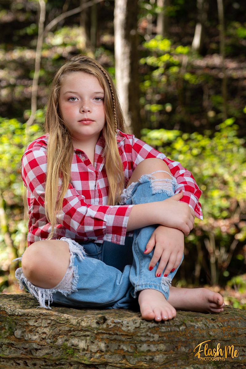 A teenage girl in a red plaid shirt and ripped jeans sits barefoot on a log in a forest.